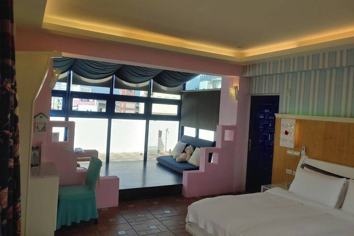 A hotel room with a bed, a seating area with a sofa and chairs, a large window with curtains, and a view of the outside.