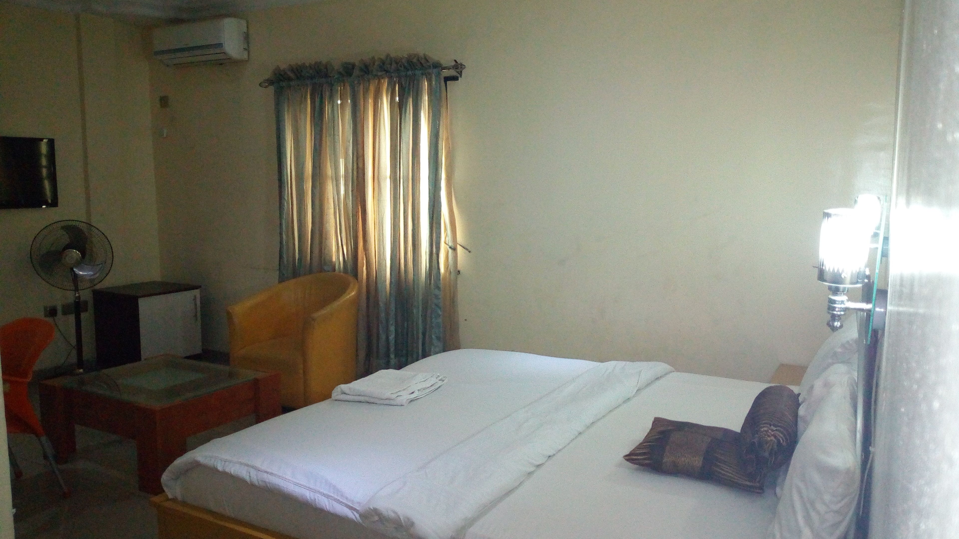 Une chambre d’hôtel avec un lit, une chaise, un bureau, un ventilateur et une fenêtre avec des rideaux.
