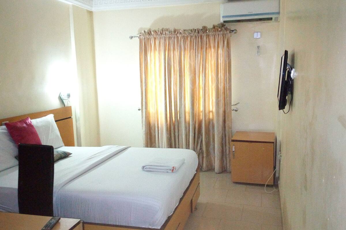 A hotel room with a bed, a small cabinet, and a curtain.