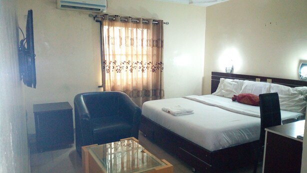 A hotel room with a bed, a desk, a chair, a glass-top coffee table, and a curtain with a floral pattern. - D'Czars Hotel & Suites (Lagos)