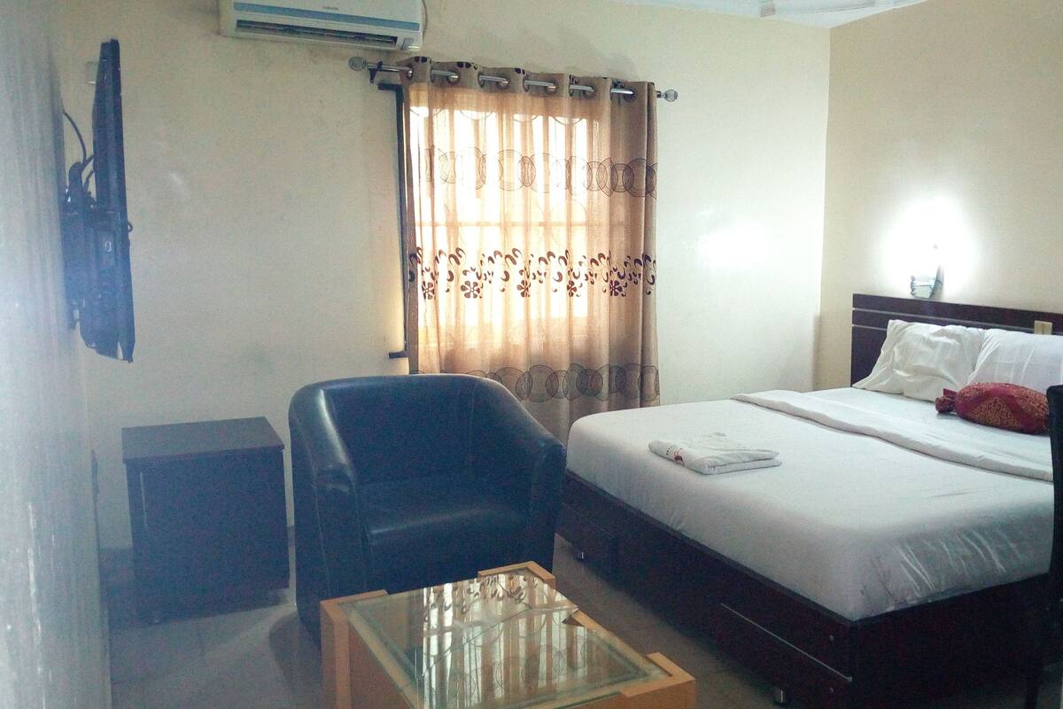 A hotel room with a bed, a desk, a chair, a glass-top coffee table, and a curtain with a floral pattern.