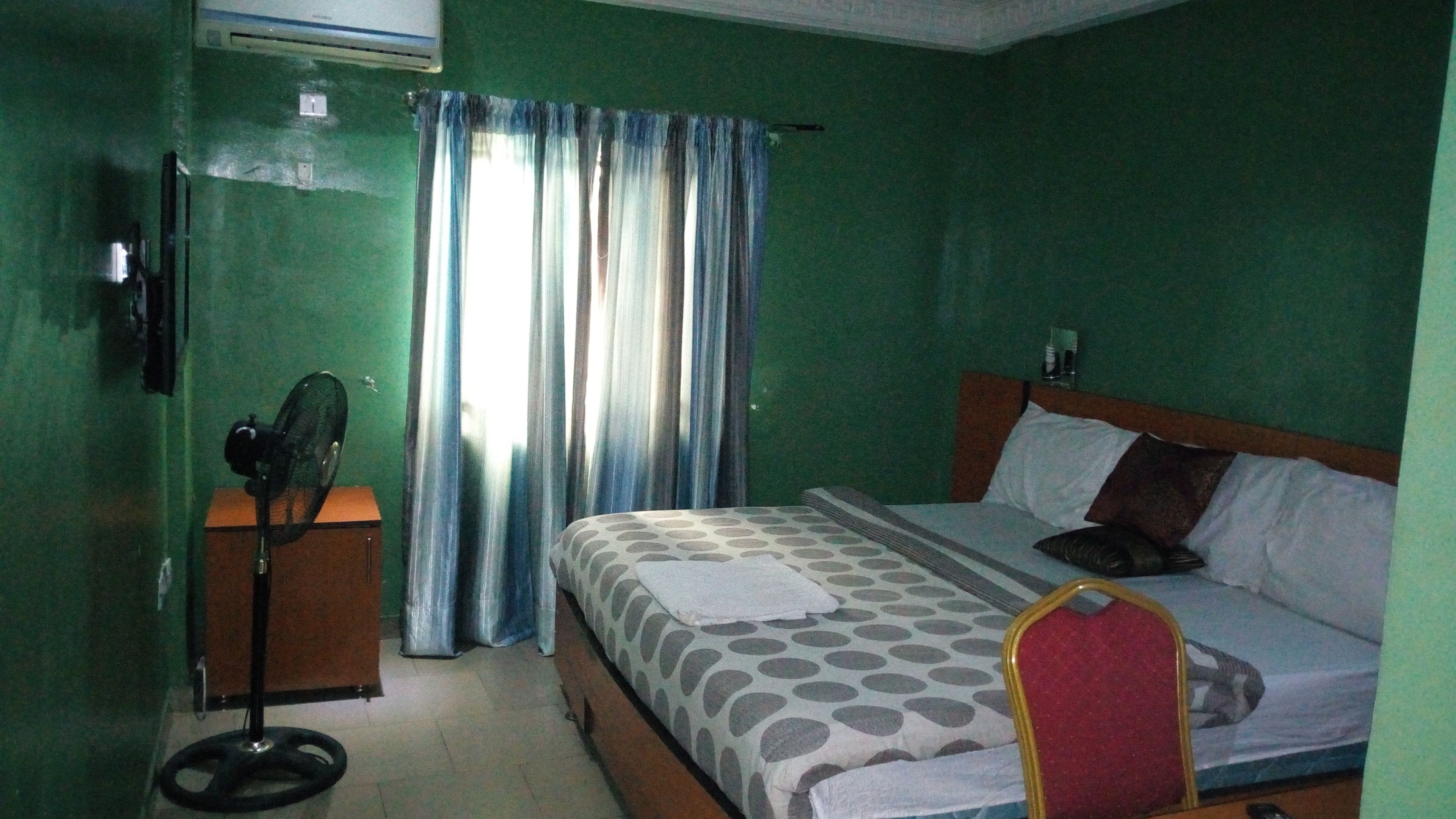 A hotel room with a bed, a desk with a fan, a window with curtains, and a wall-mounted air conditioner.