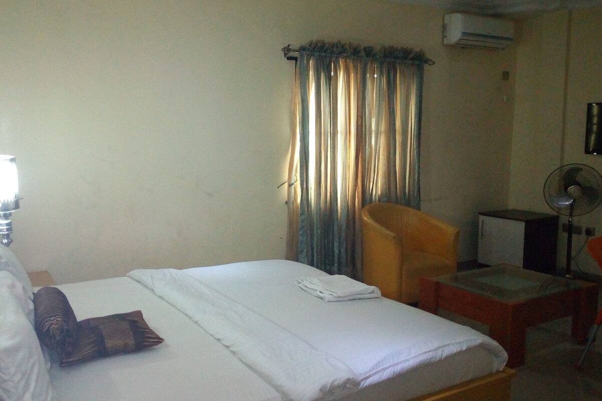 A hotel room with a bed, a chair, a desk, and an air conditioning unit.