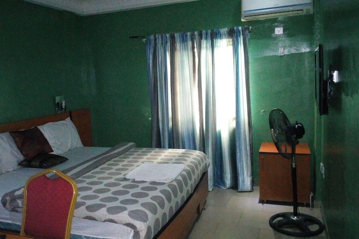 A hotel room with a bed, a bedside table with a fan, and a window with curtains.