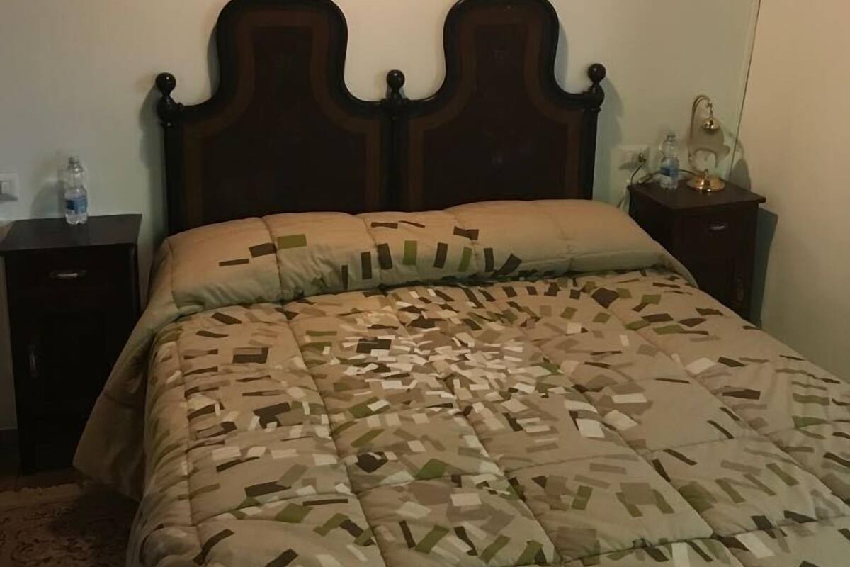 A bedroom with a wooden headboard, a patterned bedspread, a bedside table with a lamp, and a water bottle on the nightstand.