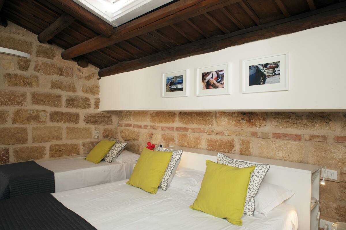 A room with two beds, each with white bedding and green pillows, set against a stone wall with wooden beams. There are three framed pictures on the wall above the beds.