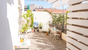 Un petit espace extérieur aménagé en patio, avec une table et des chaises, entouré de plantes en pot et d’une clôture blanche.