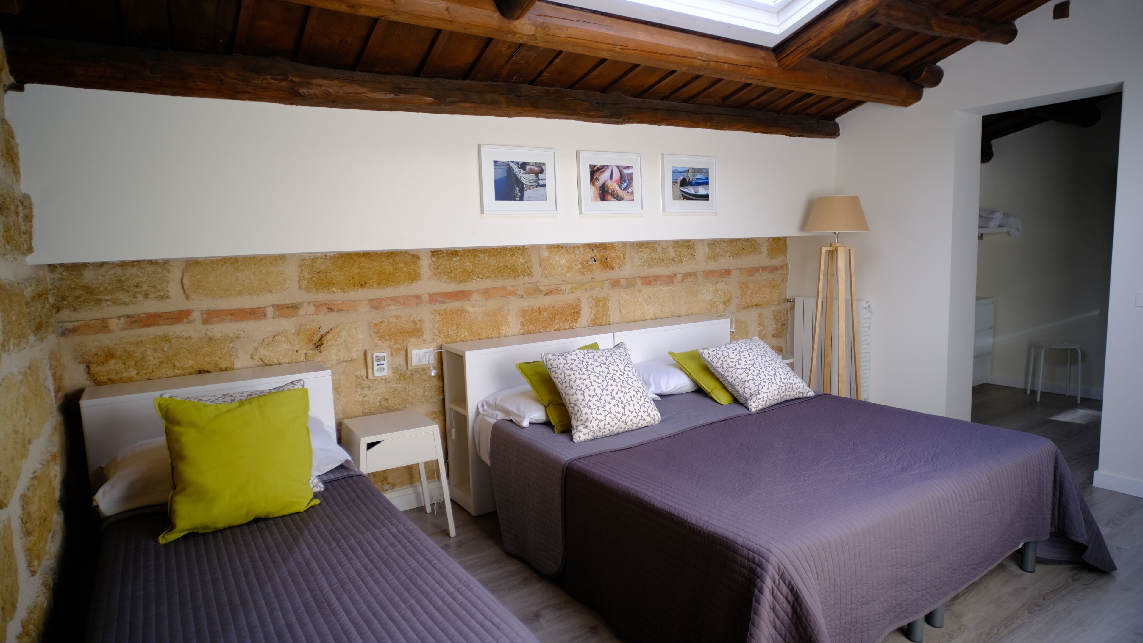 A bedroom with two beds, a brick wall, a wooden ceiling, and a small table with a lamp.