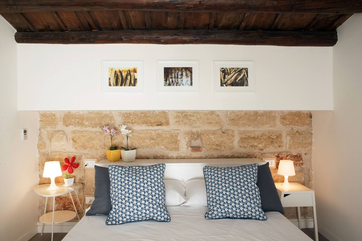 A bedroom with a stone headboard, a bed with patterned pillows, two bedside tables with lamps, and three framed pictures on the wall.
