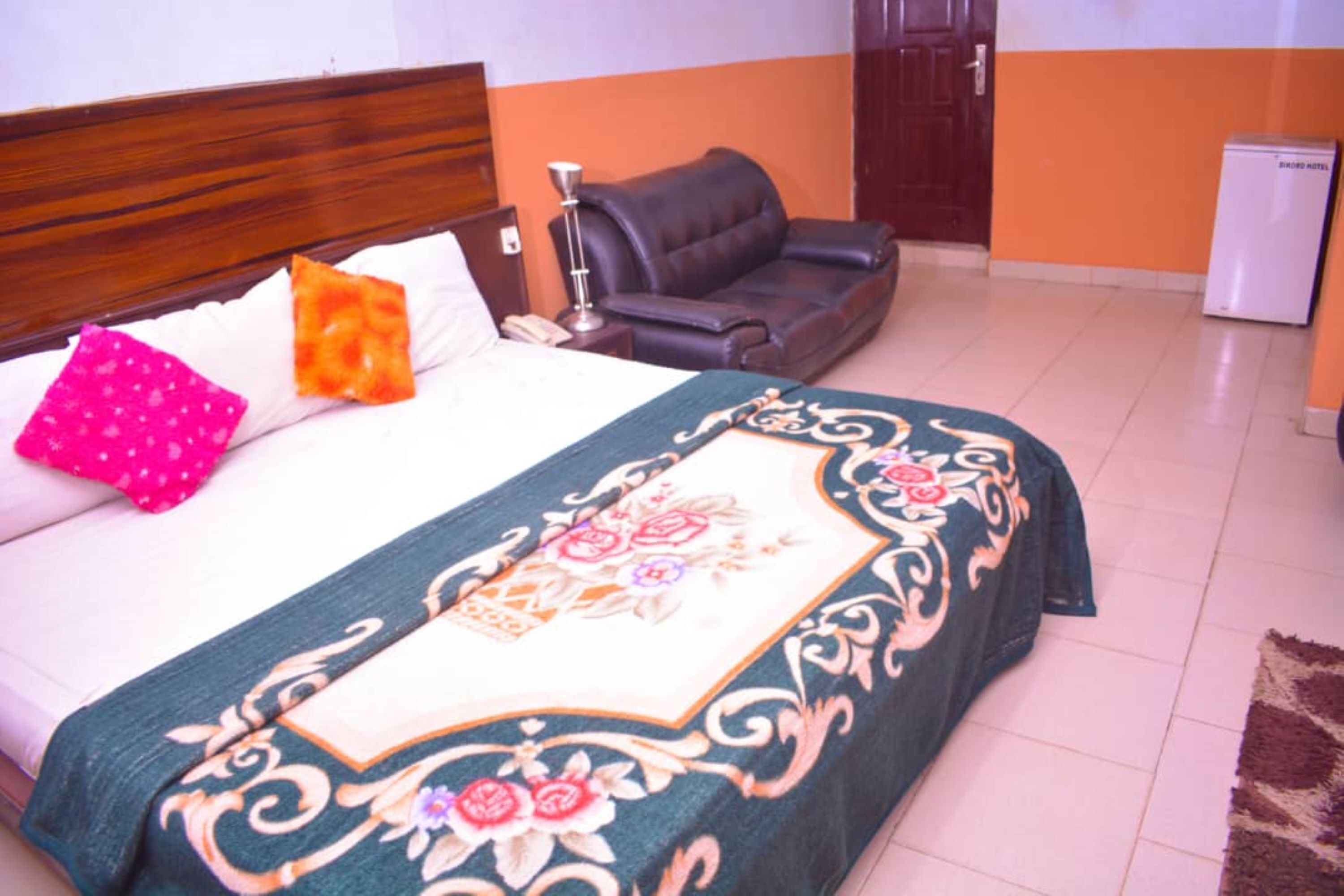 A bedroom with a bed, a wooden headboard, a black leather sofa, a small refrigerator, and a door in the background.