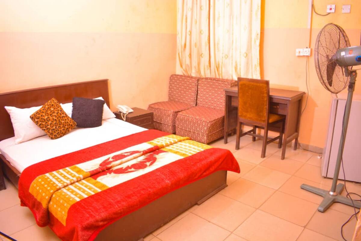 Une chambre d’hôtel avec un lit, un bureau, une chaise, un ventilateur sur pied et un rideau.