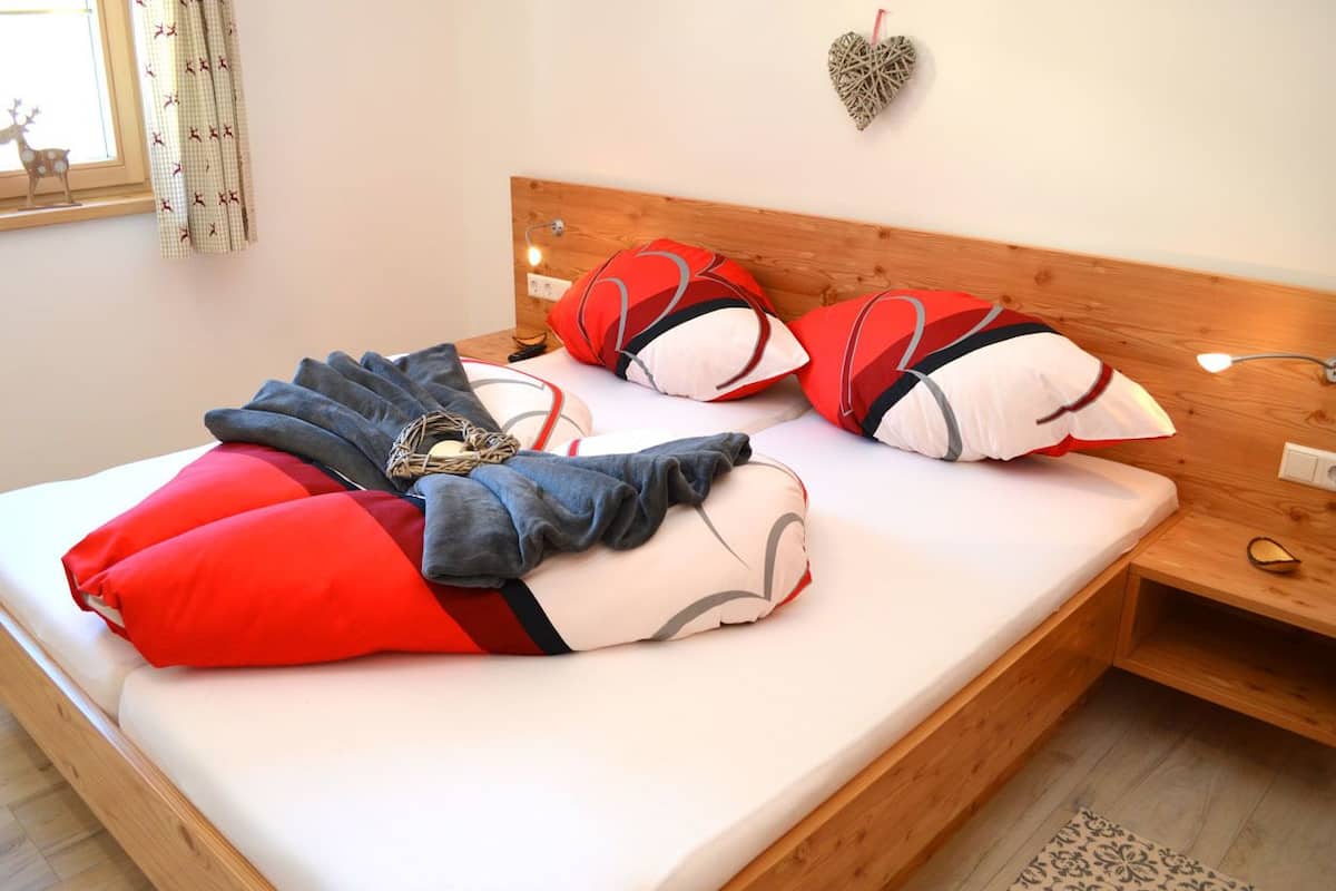 A neatly made bed with red and grey pillows, a blanket, and a small wooden nightstand with a lamp.