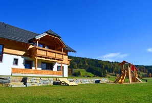 A two-story wooden house with a balcony, a stone wall, and a playground. - Denggnhof (Weisspriach)
