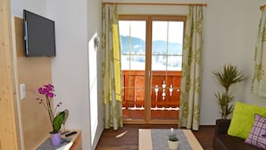 Una habitación con un escritorio de madera, televisión montada en la pared, una puerta corredera de cristal con vistas a colinas nevadas, un sofá con cojines y una pequeña planta en maceta.