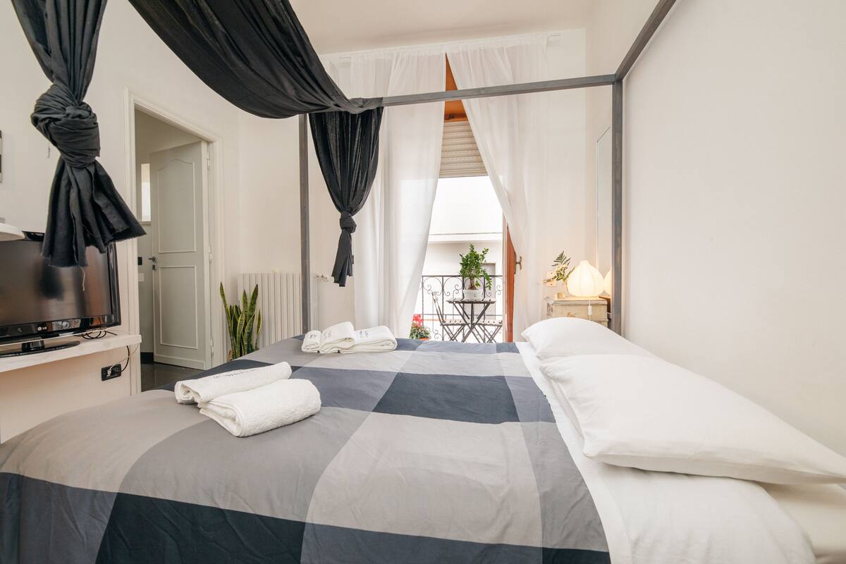 Une chambre d’hôtel avec un lit, des rideaux, une télévision, une petite table avec une plante et un balcon doté d’une rambarde.
