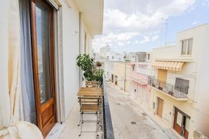 Un balcón estrecho con una mesa y sillas de madera, una planta en maceta y vistas a edificios cercanos.