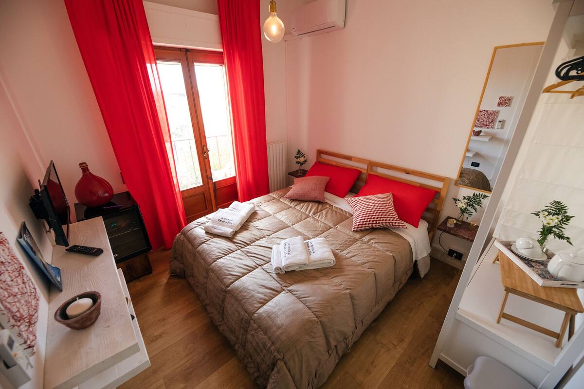 Une chambre à coucher avec un grand lit, des rideaux rouges, une table de chevet avec une lampe et une petite plante.