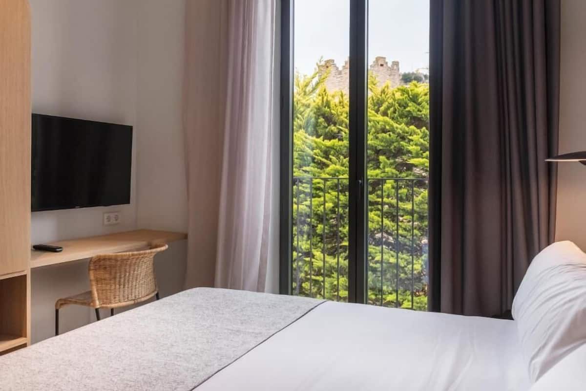 Habitación de hotel con cama, televisión, escritorio y un ventanal con vistas a zonas verdes y un edificio.