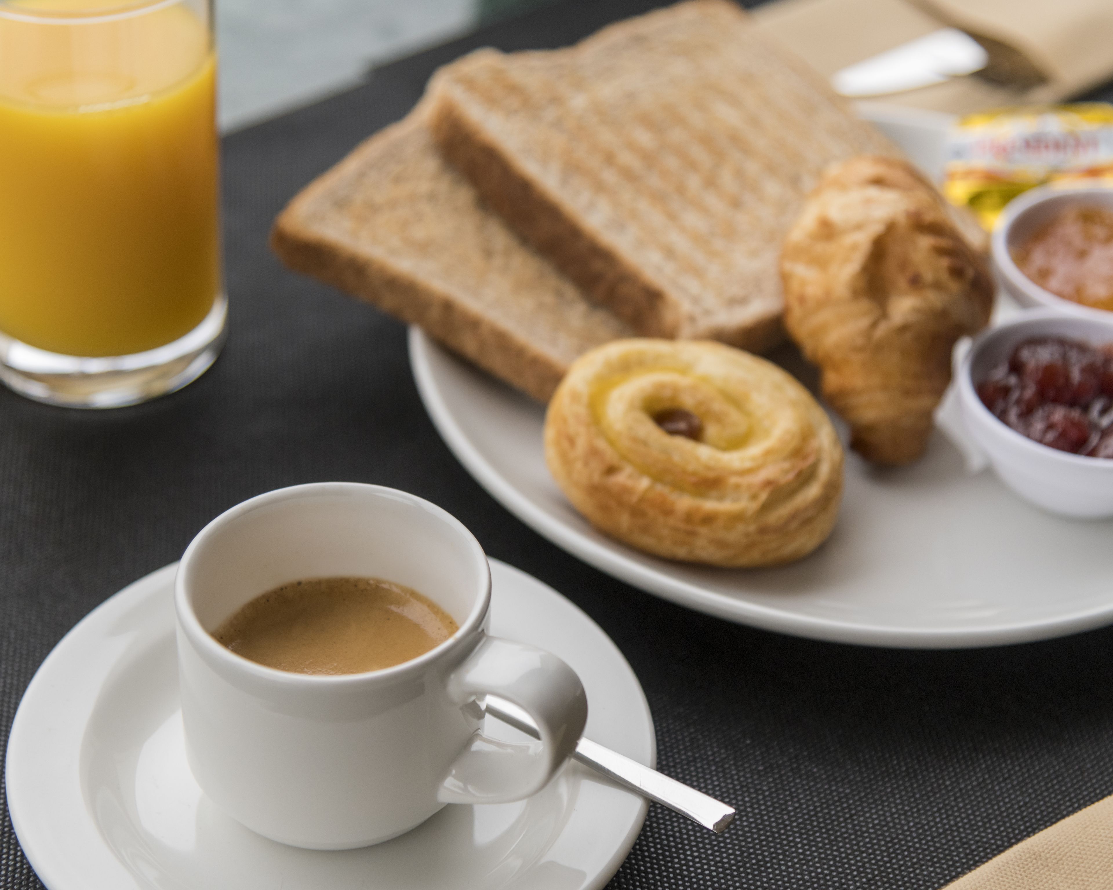 Un desayuno con una taza de café, un vaso de zumo de naranja, rebanadas de pan, un croissant y un recipiente pequeño de mermelada.