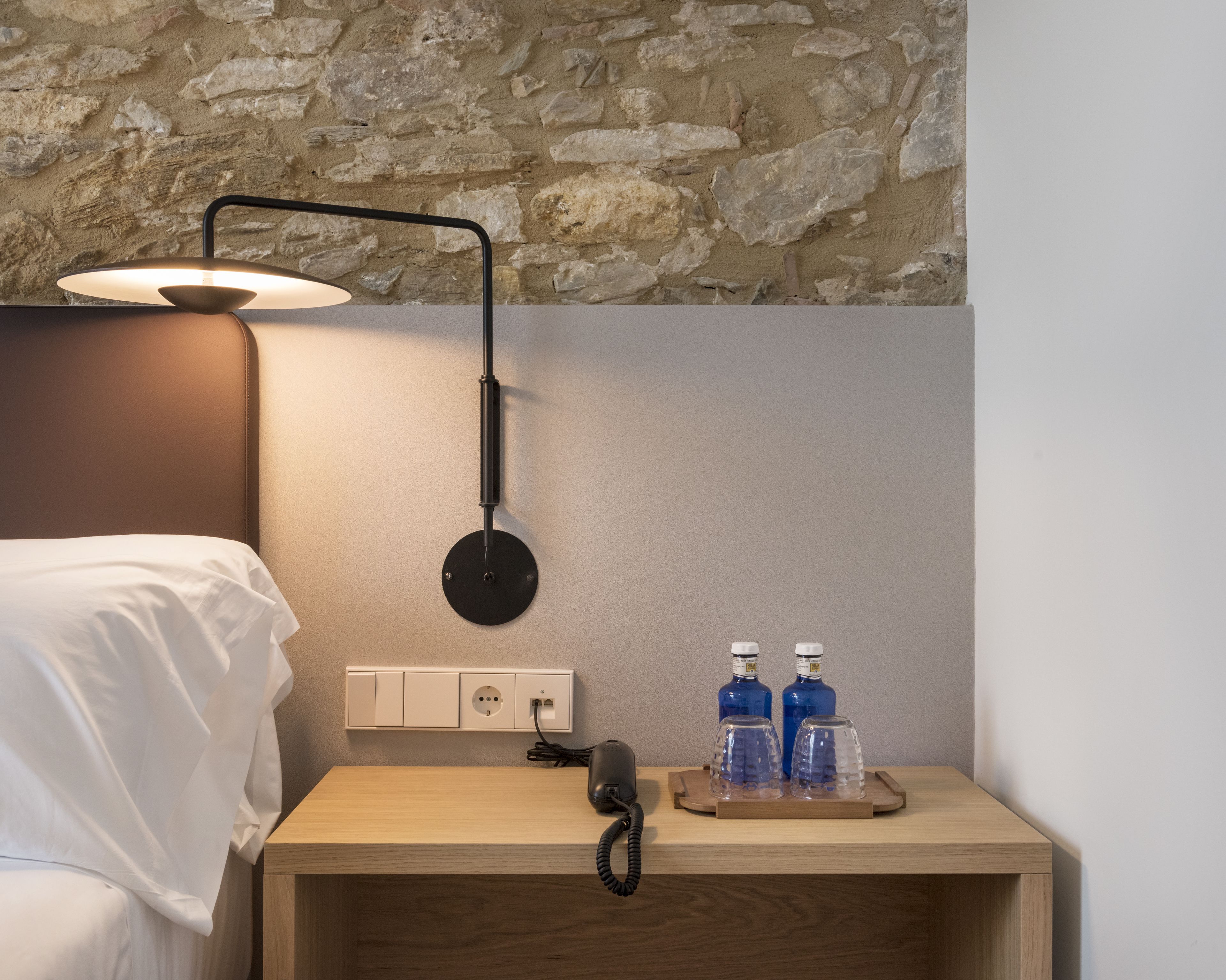 A hotel room with a bed, a bedside table with a lamp, a phone, and two bottles on the table.