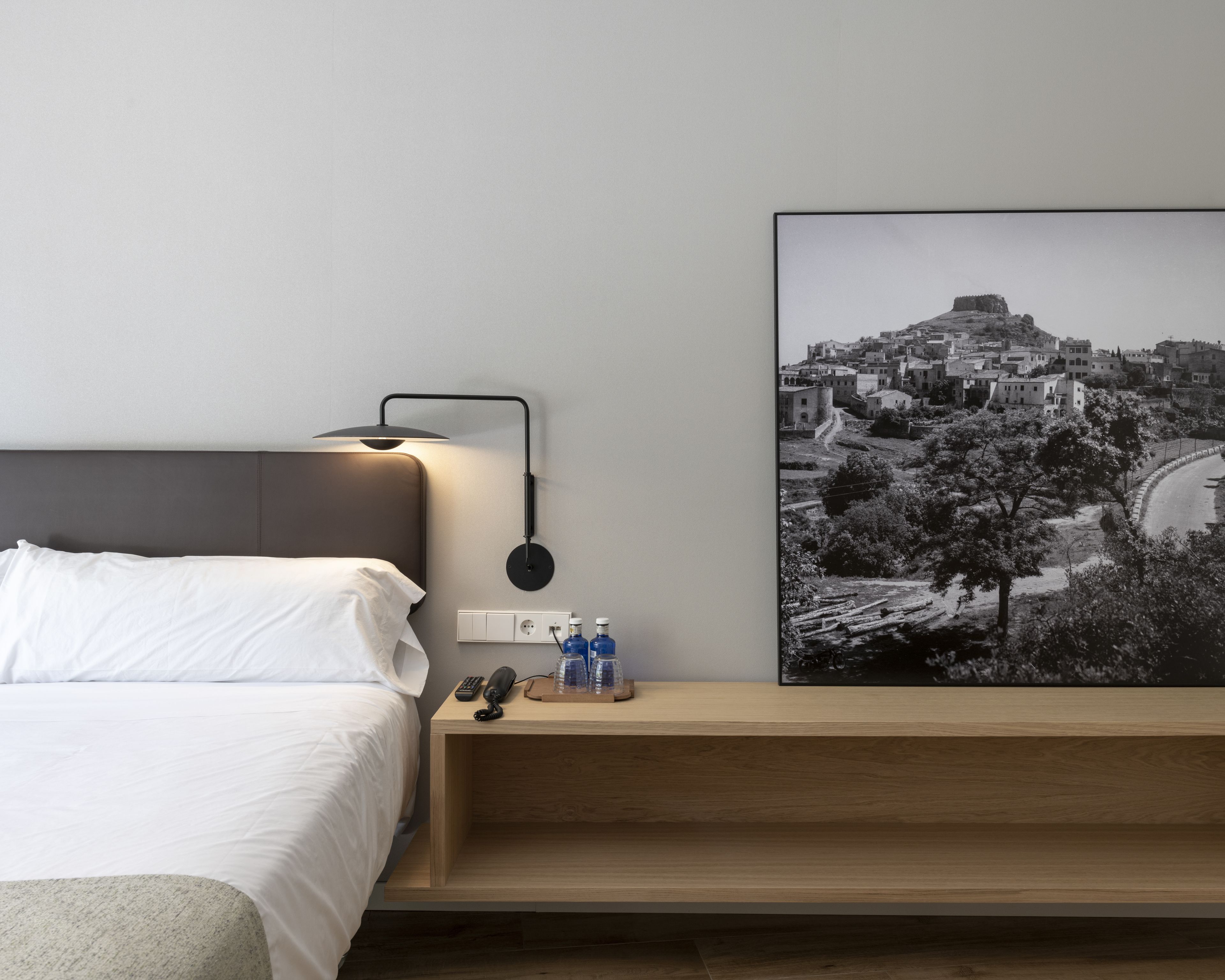 A modern hotel room with a bed, bedside table, lamp, and a framed black and white photograph of a town.