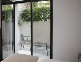 Una stanza con un letto e una porta scorrevole in vetro che dà su un patio esterno con due sedie verdi e una panca.