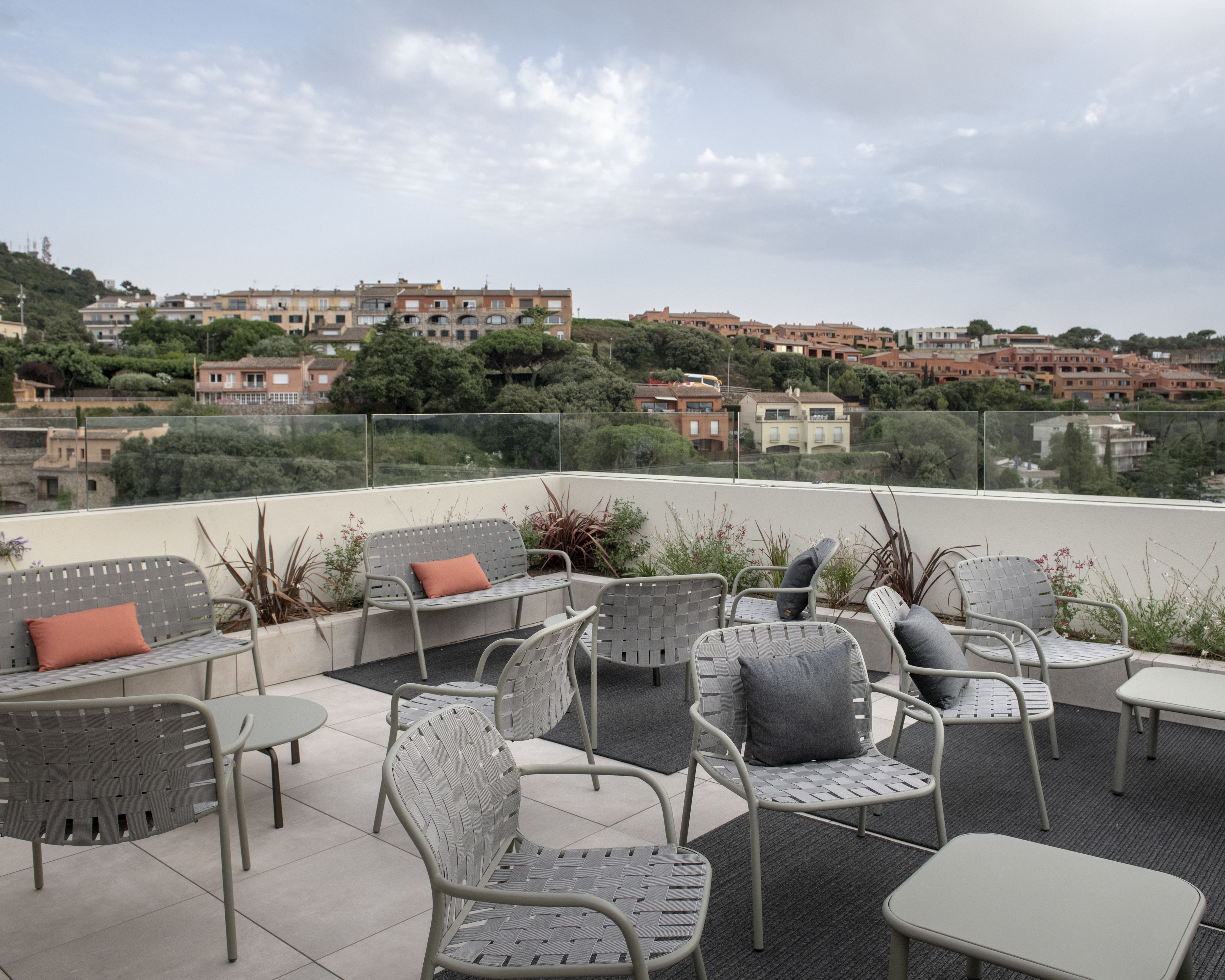 An outdoor patio area with metal chairs and tables, surrounded by a glass railing, overlooking a residential area with buildings and greenery.