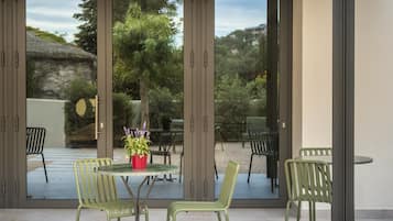 A modern outdoor dining area with green chairs and a table, large sliding glass doors, and a view of a garden and buildings.