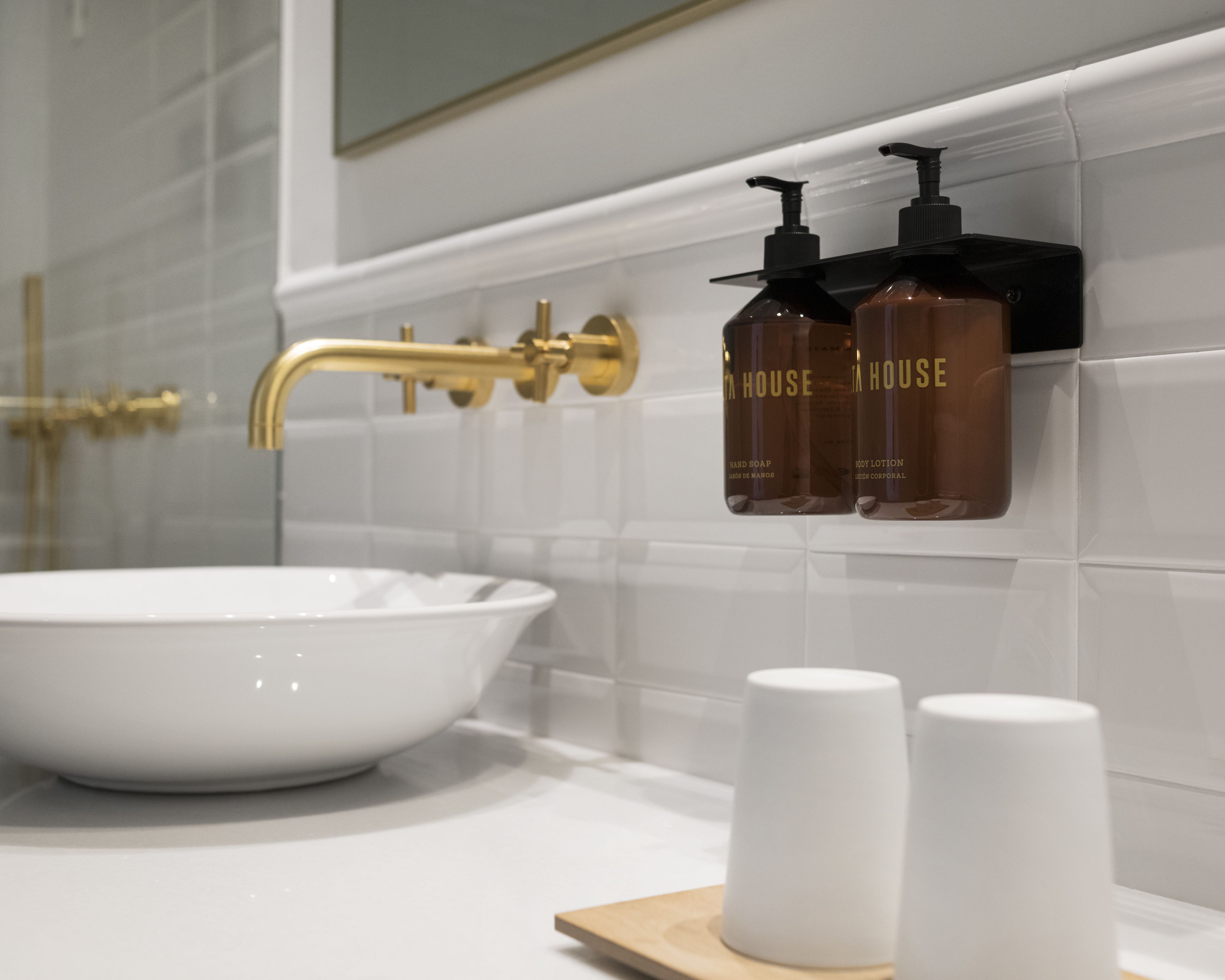Un baño con un lavabo blanco, grifo dorado y dos botellas de jabón de manos y crema corporal sobre una pared de azulejos blancos.