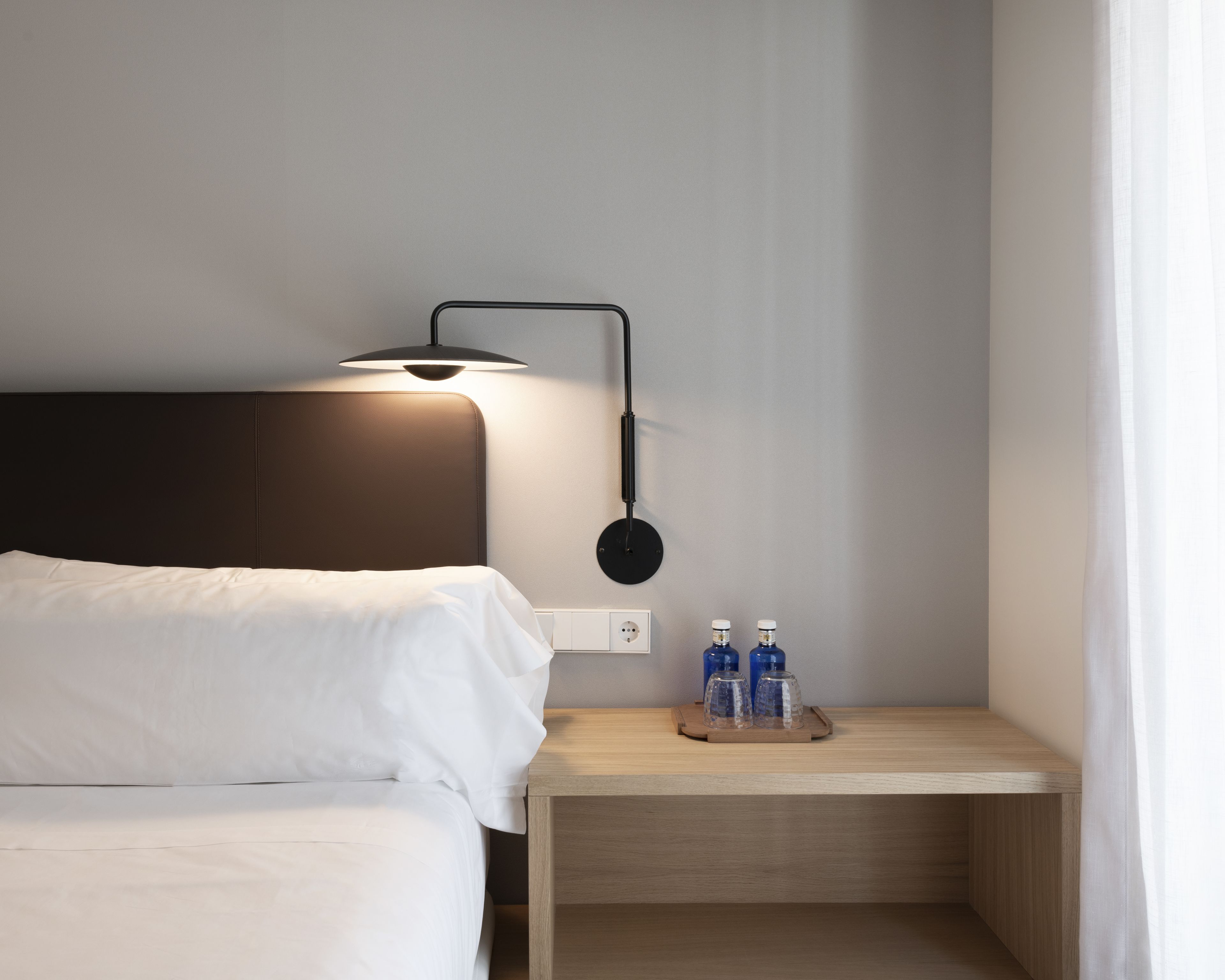 A neatly made bed with white linens, a bedside table with a lamp, and two bottles on the table.