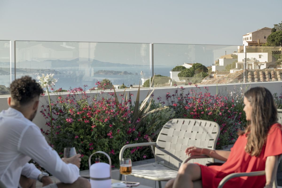 Un balcon donnant sur une ville côtière, une table avec un verre de vin et deux personnes assises.