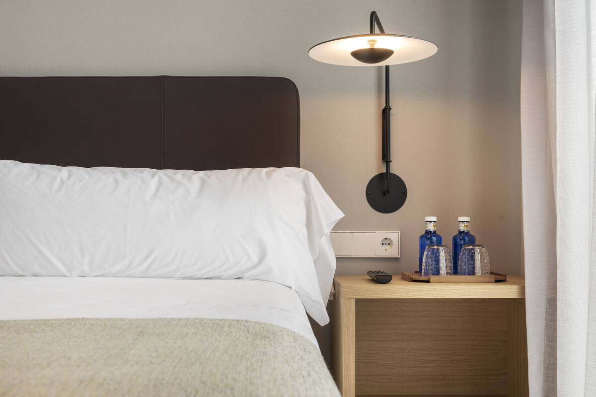 Una cama bien tendida con sábanas blancas, una mesita de noche con dos botellas y una lámpara de pared.