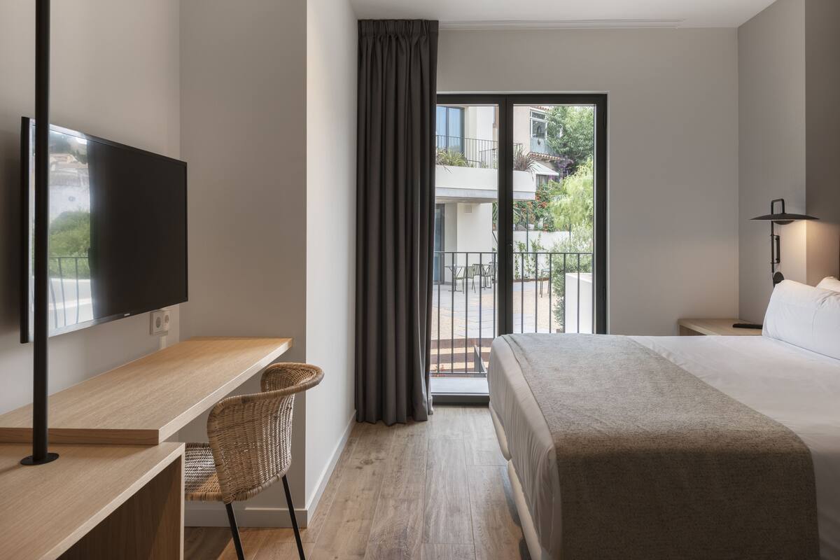 Una habitación de hotel moderna con una televisión de pantalla plana, un escritorio de madera, una silla de mimbre, una cama grande y un balcón con vistas al exterior.
