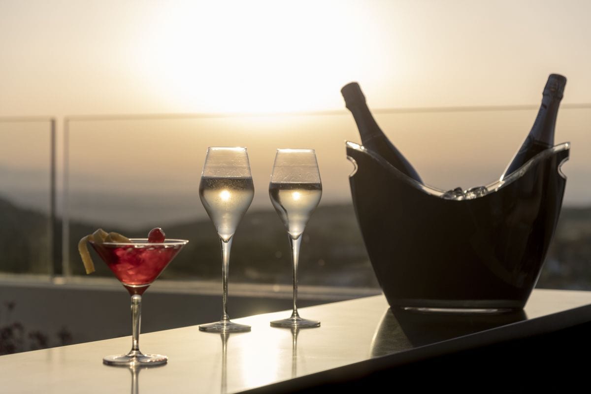 Uma mesa com duas taças de champanhe e um copo de martini, uma garrafa de champanhe e um balde de gelo, com vista para o pôr do sol.