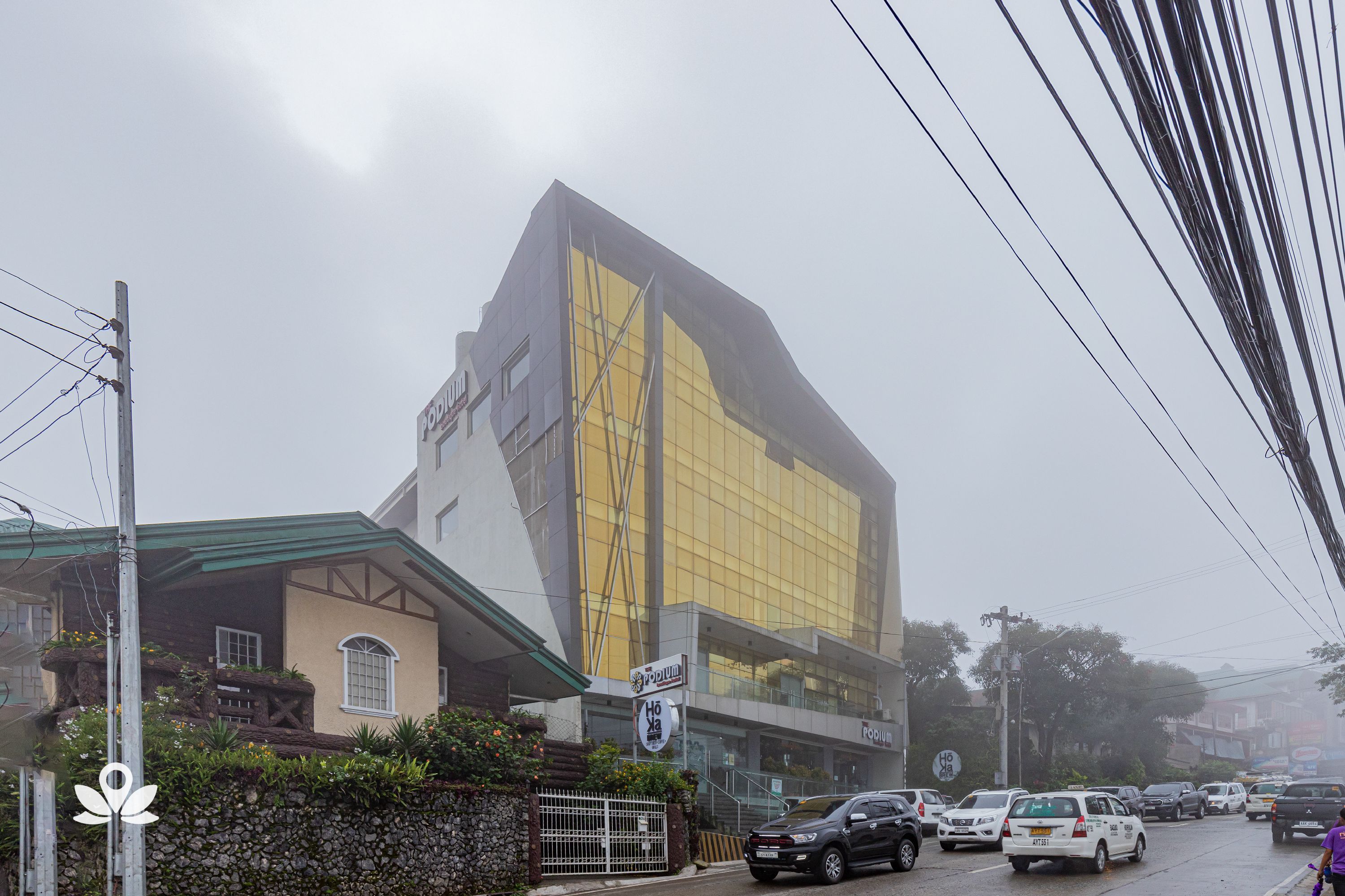 Un edificio moderno con una amplia fachada de vidrio, un muro de piedra con vegetación y una calle con coches estacionados.