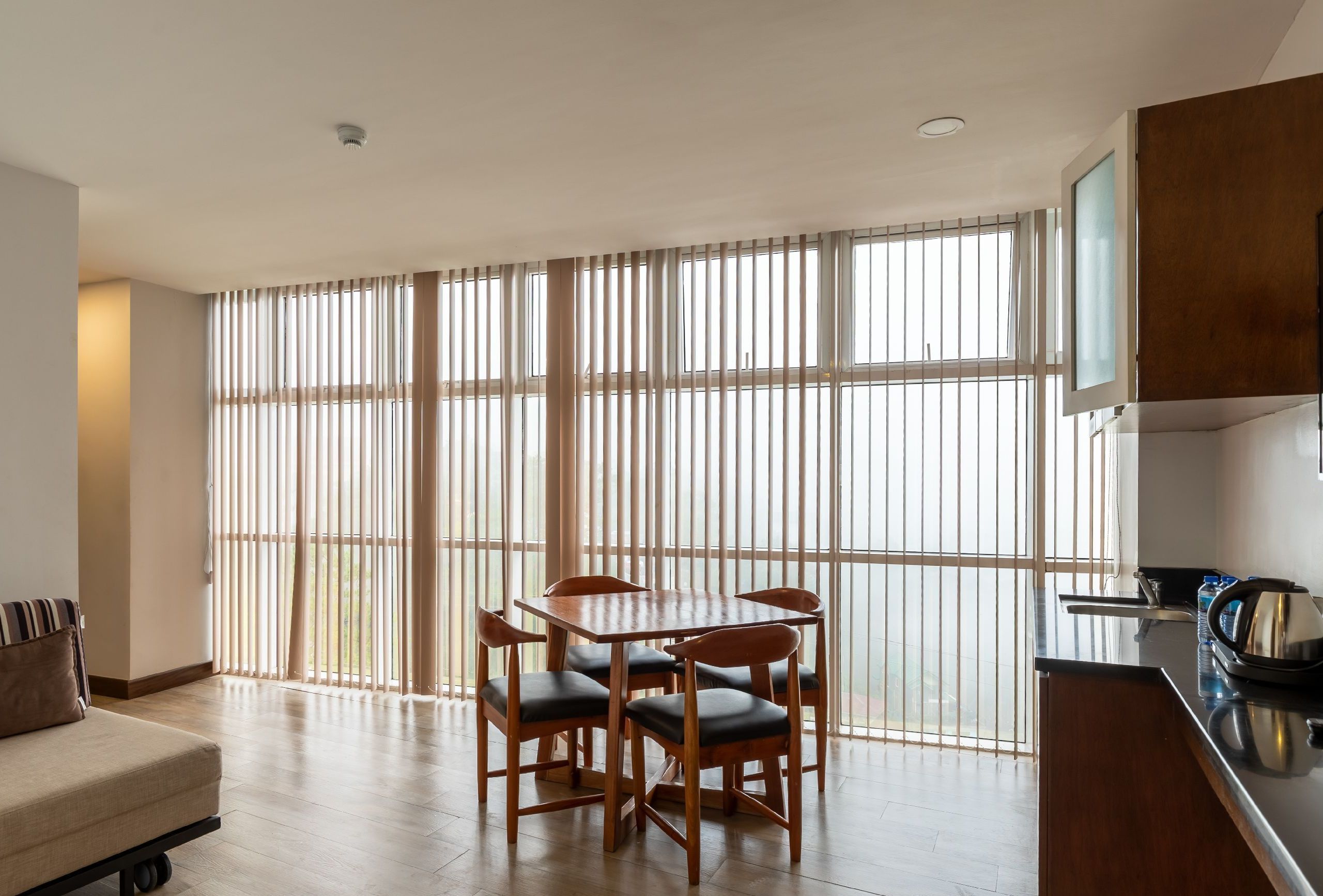 A modern kitchen with a dining area, large windows with blinds, and a wooden table with chairs.