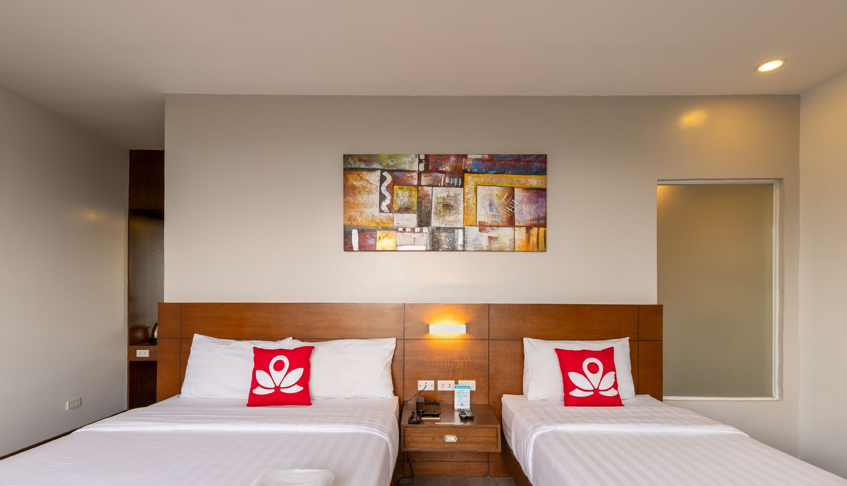 Habitación de hotel con dos camas, un cabecero de madera, una obra de arte en la pared, una mesita de noche con lámpara y un enchufe.