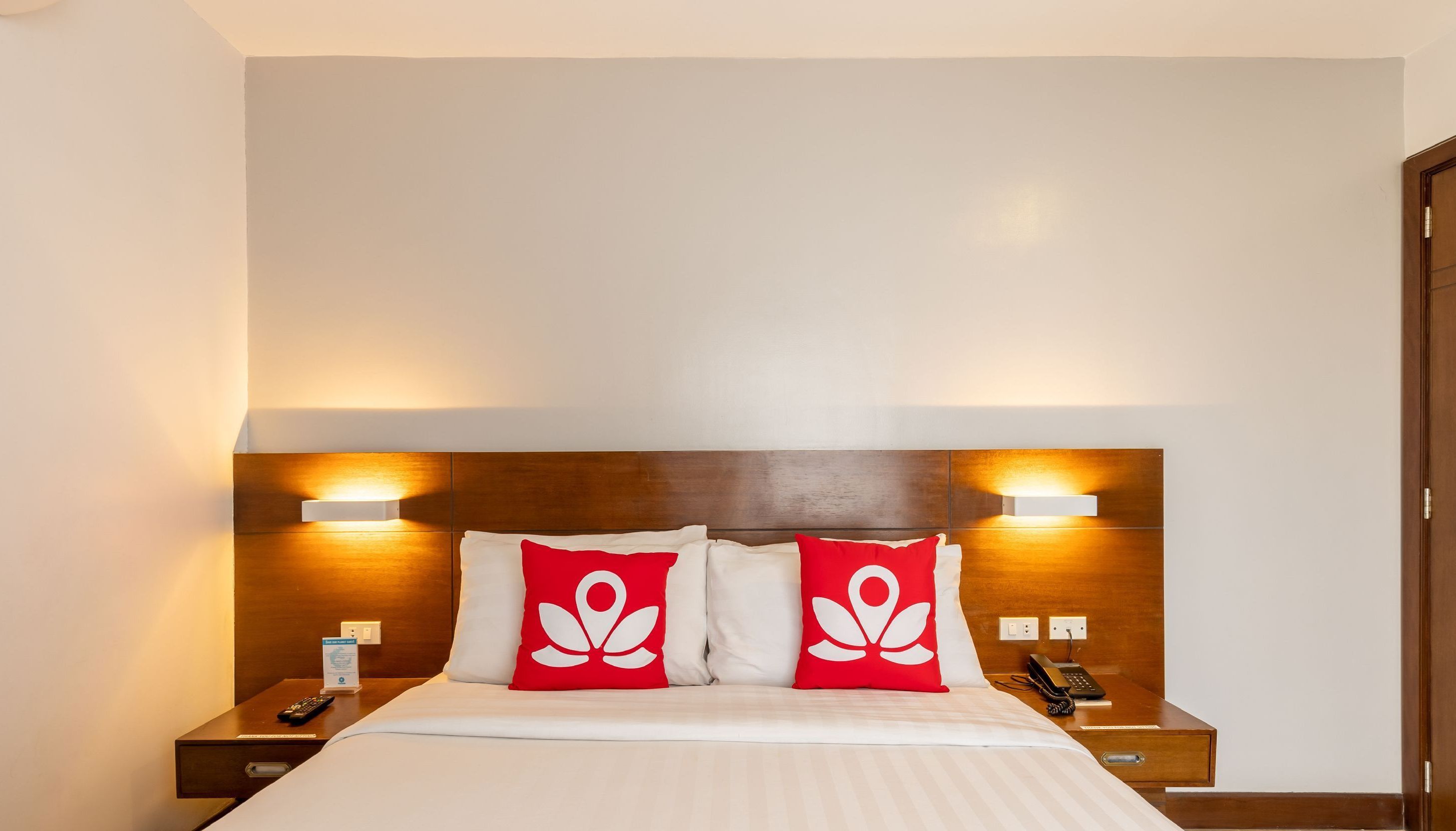Una habitación de hotel con cabecera de madera, dos mesitas de noche, una cama con ropa blanca y almohadas rojas con diseño blanco.