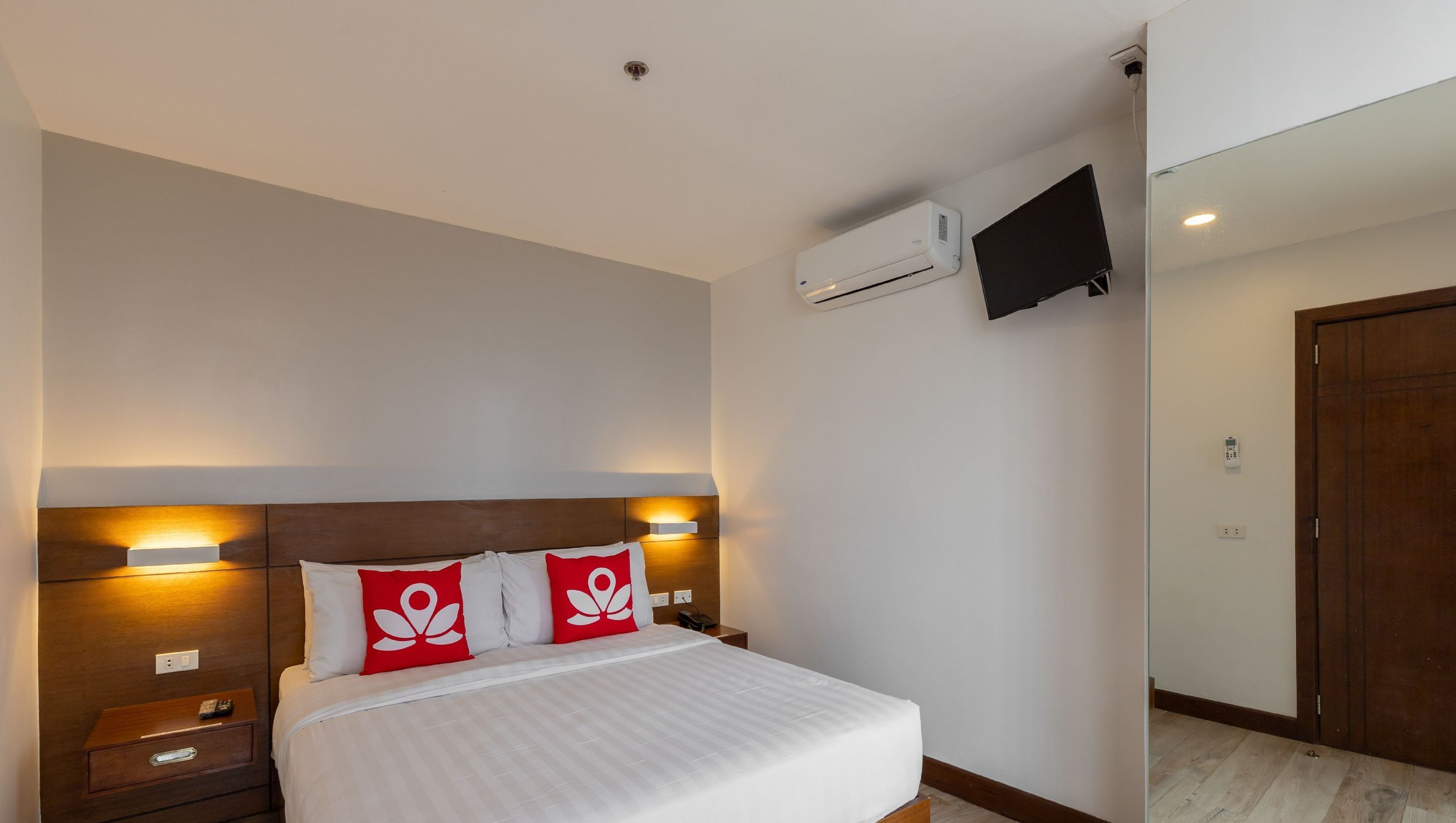 A hotel room with a bed, bedside tables, a TV mounted on the wall, and an air conditioning unit.