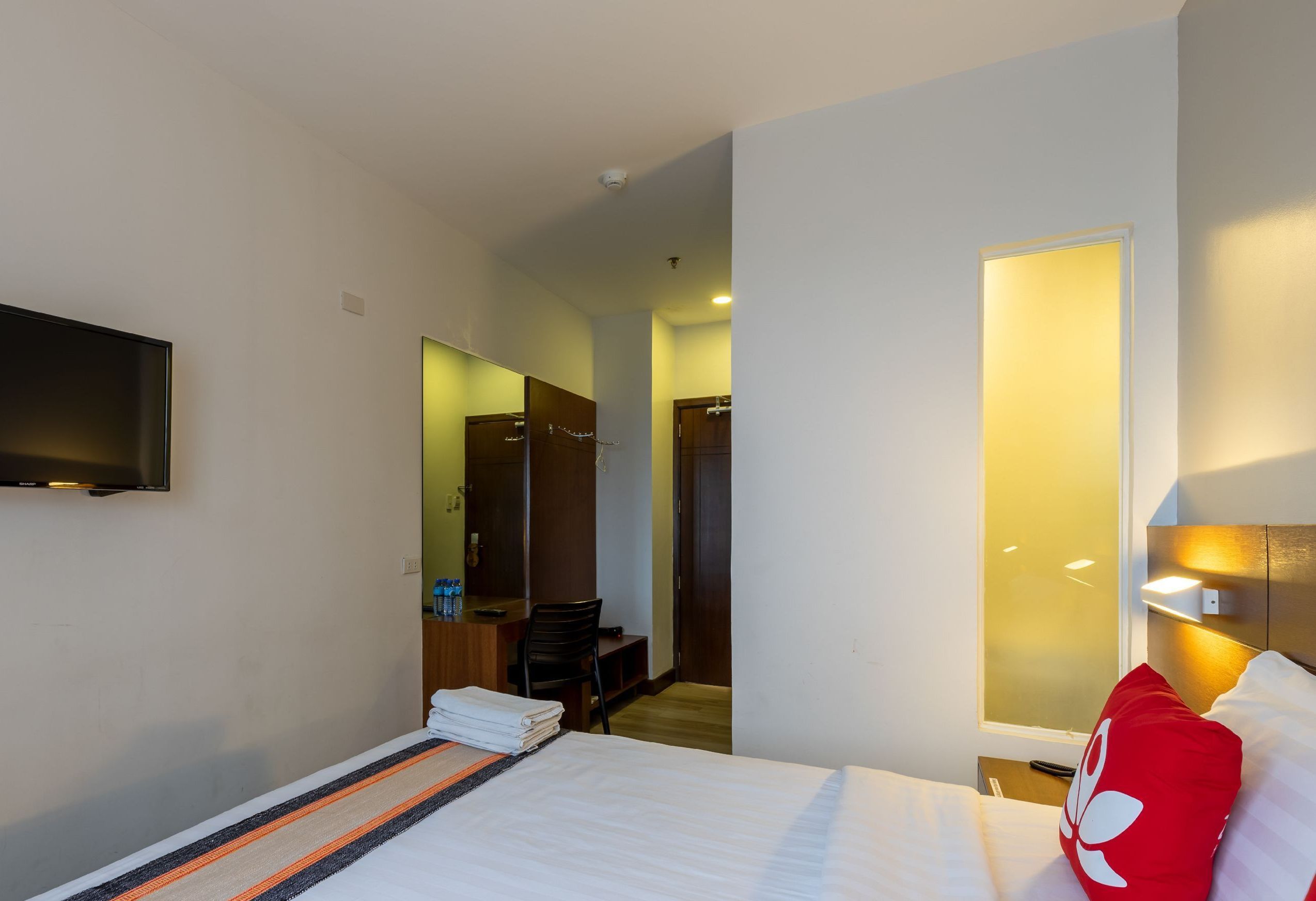 A hotel room with a bed, a TV mounted on the wall, a desk with a chair, and a bathroom visible through a glass door.