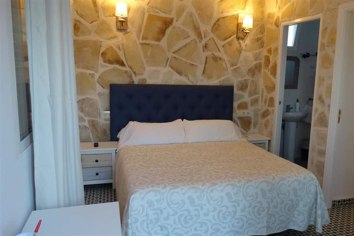 A bedroom with a stone wall, a bed with a dark headboard, bedside tables, and a bathroom visible through an open door.