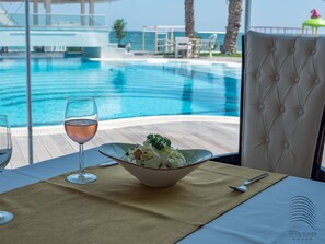 A dining table with a bowl of salad, a wine glass, and a fork. A view of a swimming pool and palm trees is visible through a window. - White Tower Mamaia (Navodari)