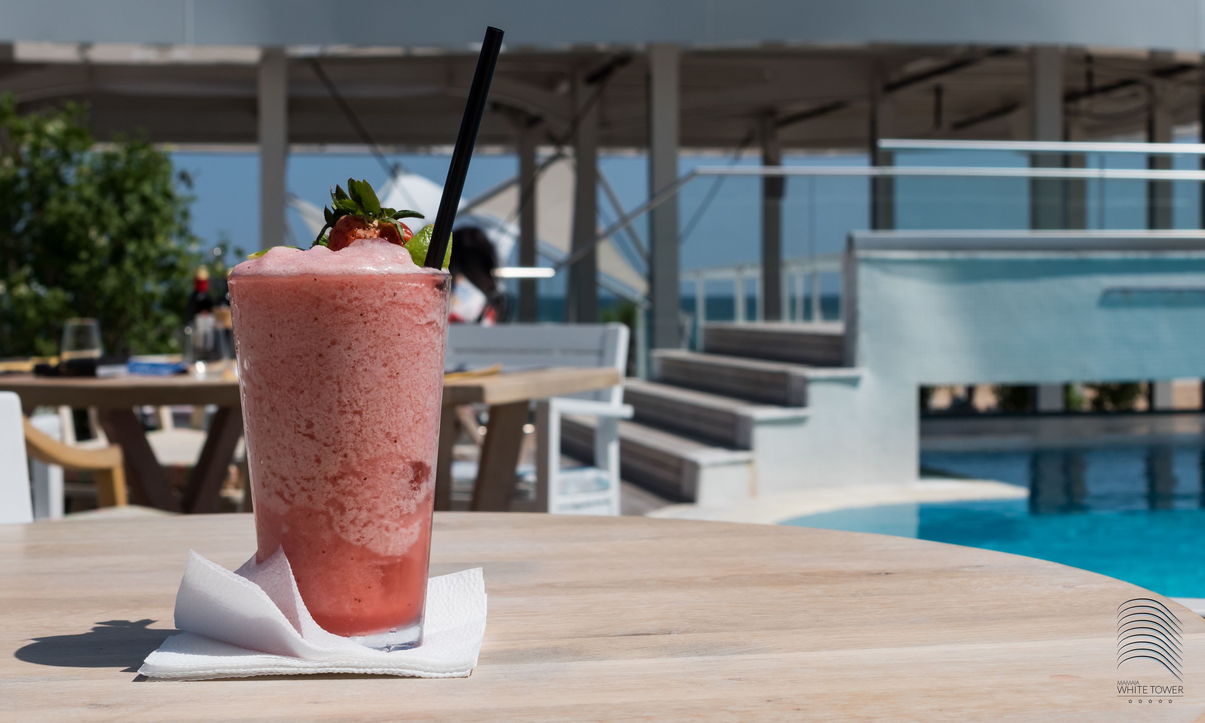 A pink smoothie on a wooden table near a poolside area with a view of a building and a pool.