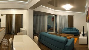 A modern living room with a sofa, a coffee table, and a TV. There is a dining area with a table and chairs. The room has a large window with curtains. - White Tower Mamaia (Navodari)