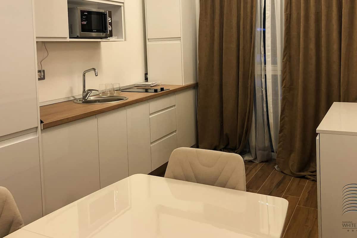 A compact kitchen with a microwave, sink, and wooden countertop. There is a dining table with chairs and a view of a window with curtains.