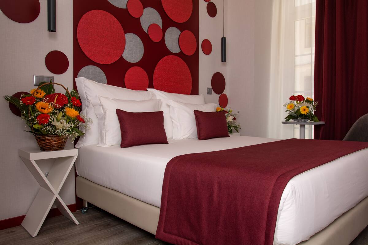 A neatly made bed with a red blanket, a white pillow arrangement, and a side table with a basket of flowers.
