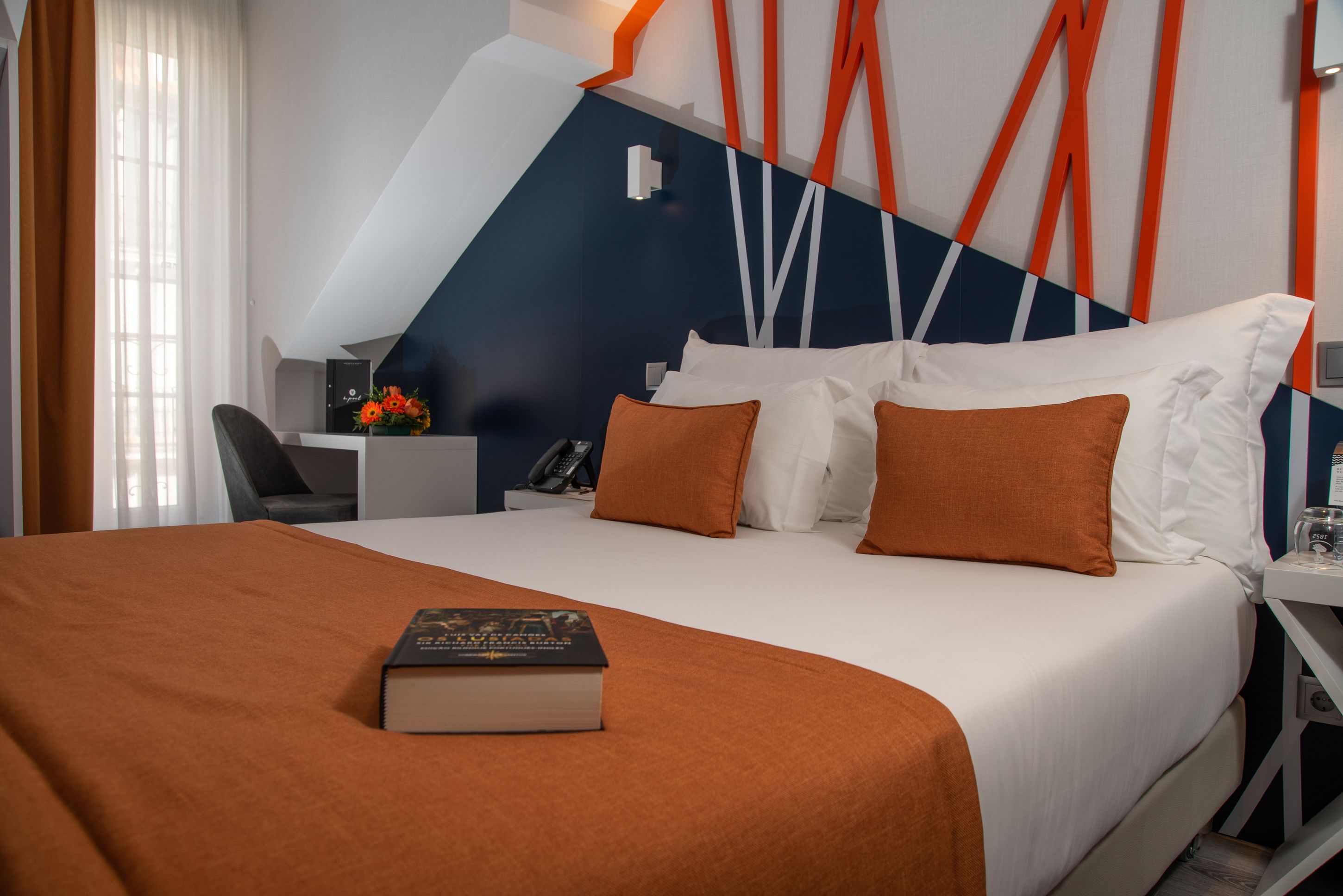 Une chambre d’hôtel avec un grand lit, un bureau et une chaise. Il y a un livre sur le lit et un vase rempli de fleurs sur le bureau.