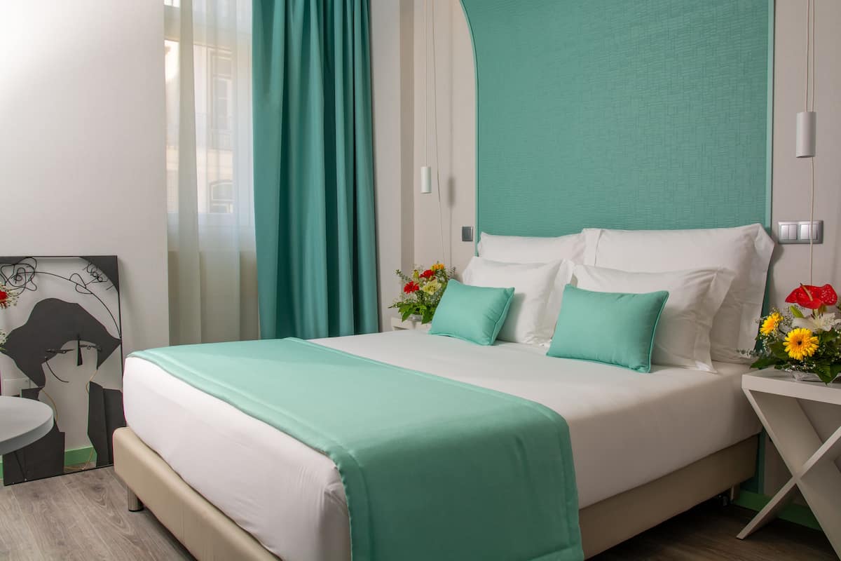 A neatly made bed with white and teal bedding, a teal headboard, and a small bedside table with a vase of flowers.