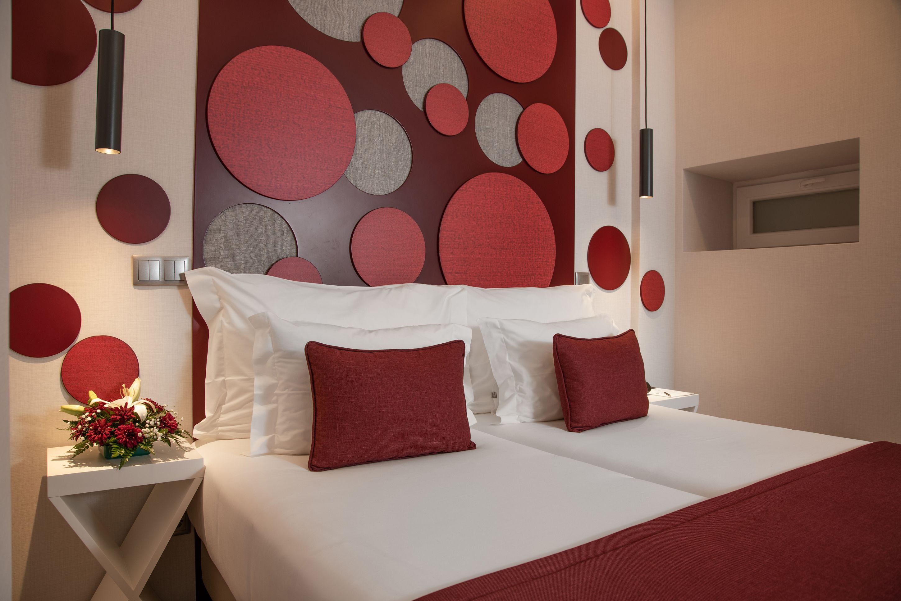 A modern hotel room with a large bed, red and grey circular wall decor, white bedding, and a bedside table with a vase of flowers.