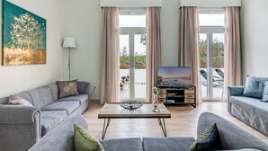 Un soggiorno moderno con divani grigi, un tavolino da caffè e una TV. Ampie finestre con tende offrono luce naturale e vista sull'esterno.